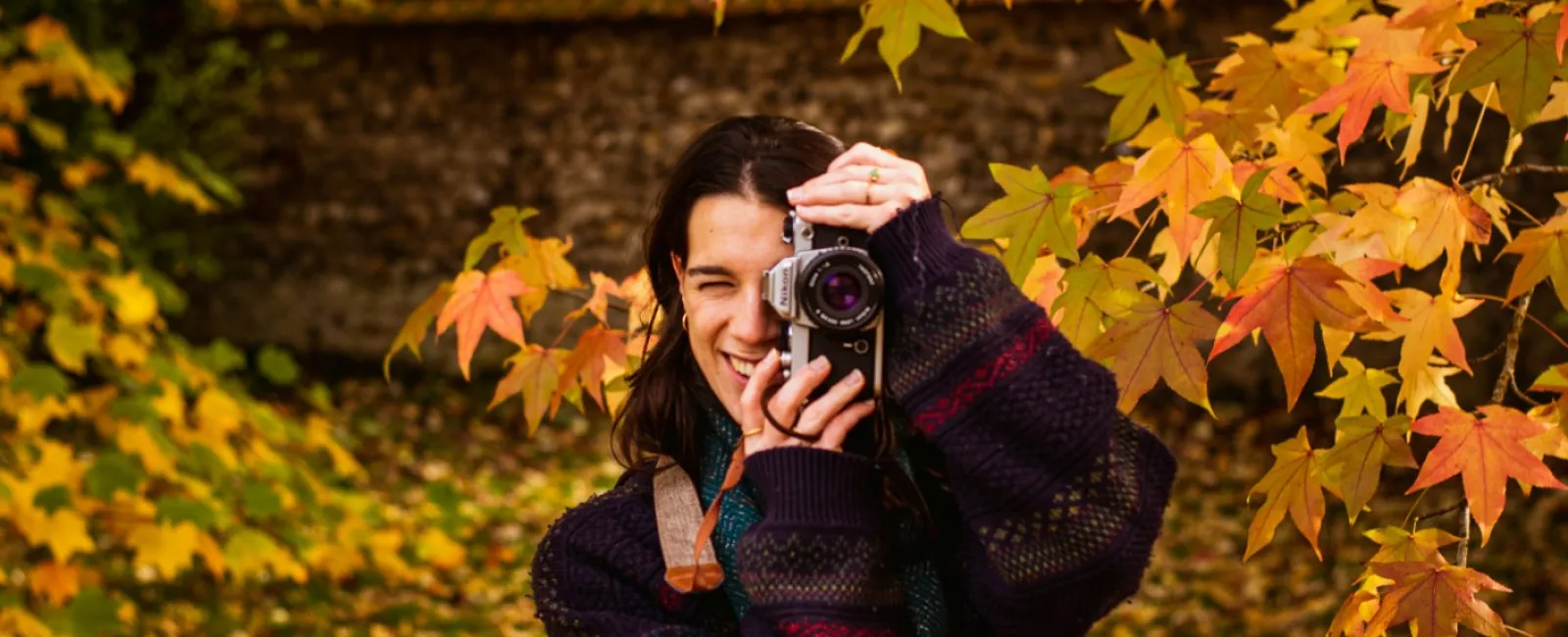 Woman in autumn sweater holding a camera amidst vibrant fall leaves in a forest setting.