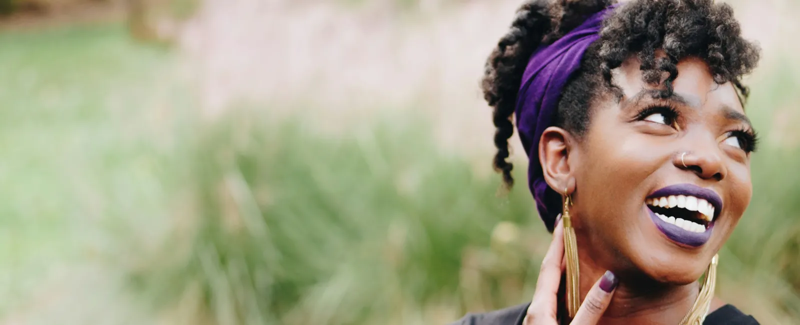 Smiling woman with curly hair wearing purple headband and lipstick, large earrings, and gold watch outdoors.