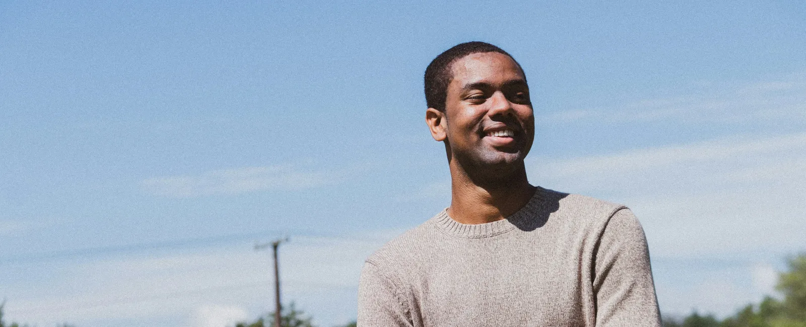 Smiling man wearing a beige sweater standing with arms crossed in a sunflower field under a blue sky.