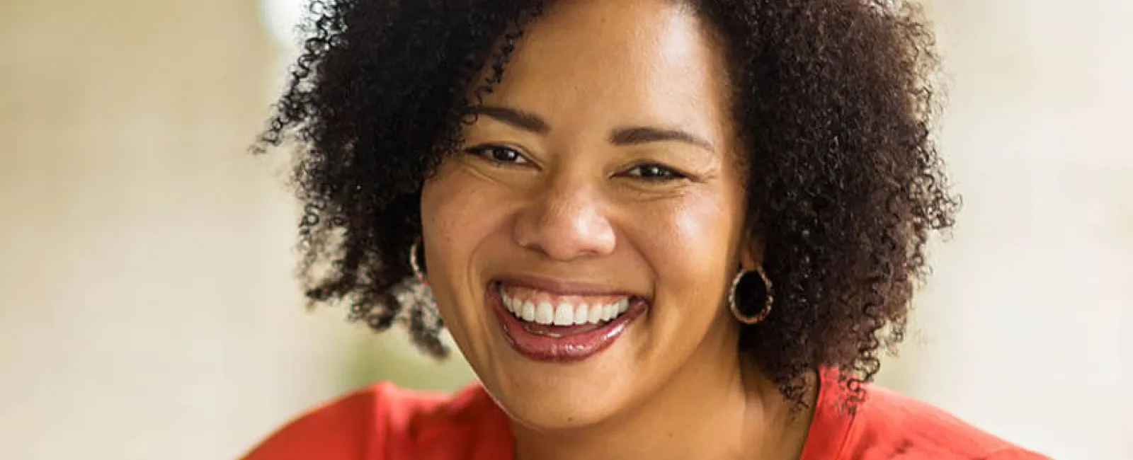 Cheerful woman with curly hair smiling warmly, wearing a vibrant orange blouse in a natural outdoor setting.