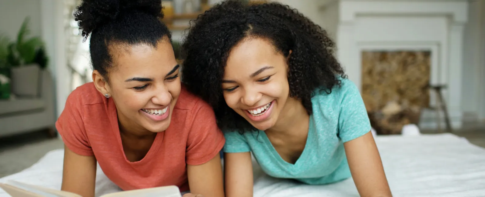 Two smiling teenage girls lying on bed reading a book and using a tablet together in a cozy room.