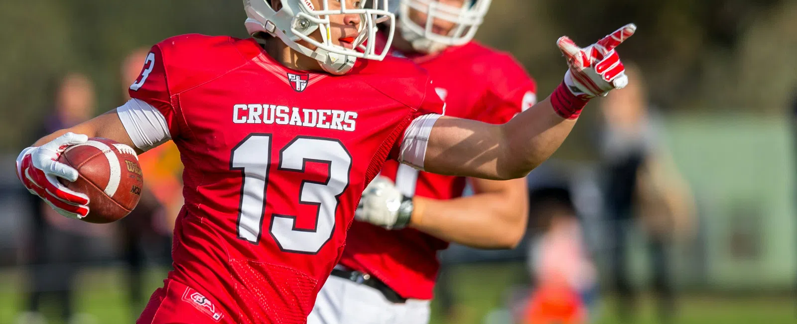 Football player in red Crusaders uniform with number 13 running with ball and pointing during game