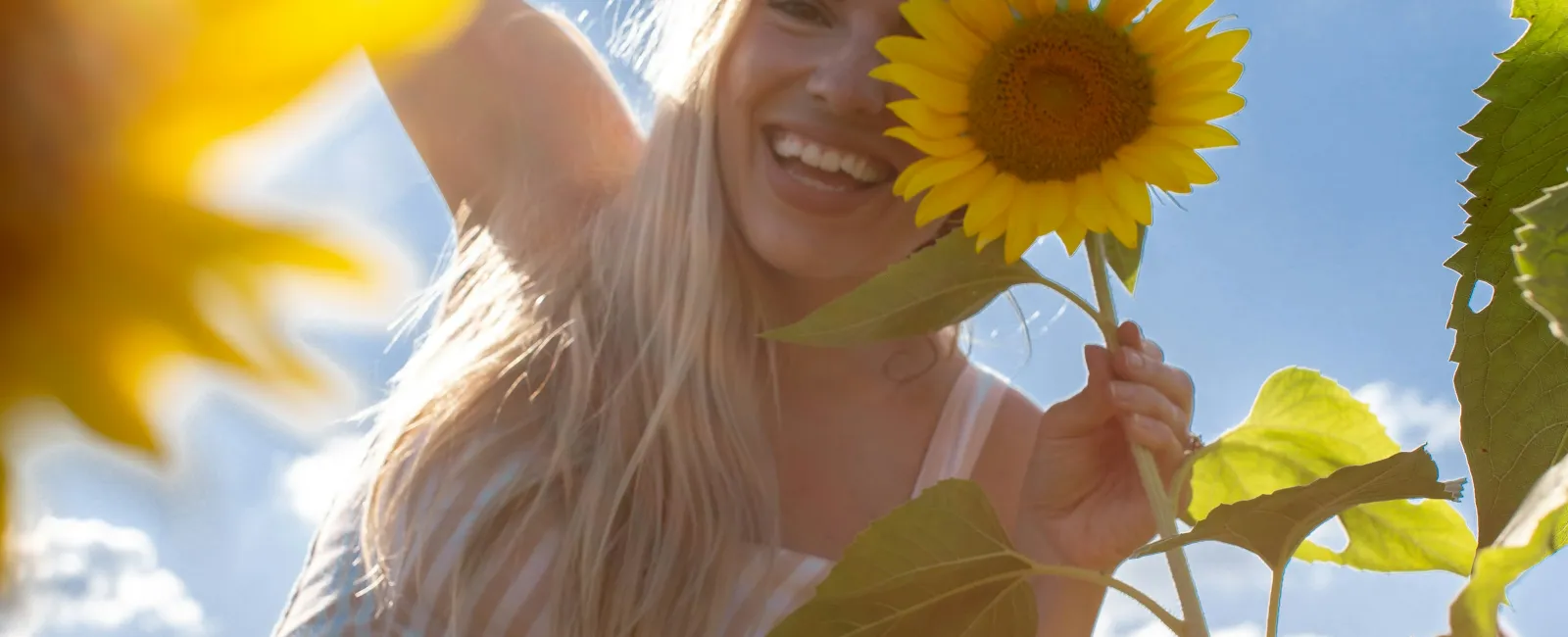Smiling woman holding a sunflower in a field under blue sky with clouds, wearing striped dress with buttons.