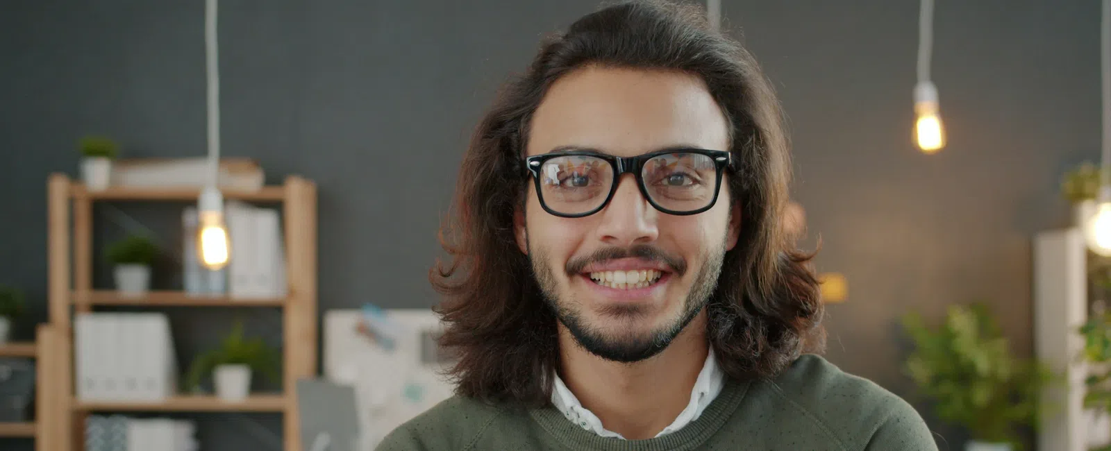 Smiling man with glasses and long hair wearing a green sweater in a modern workspace with plants and shelves.