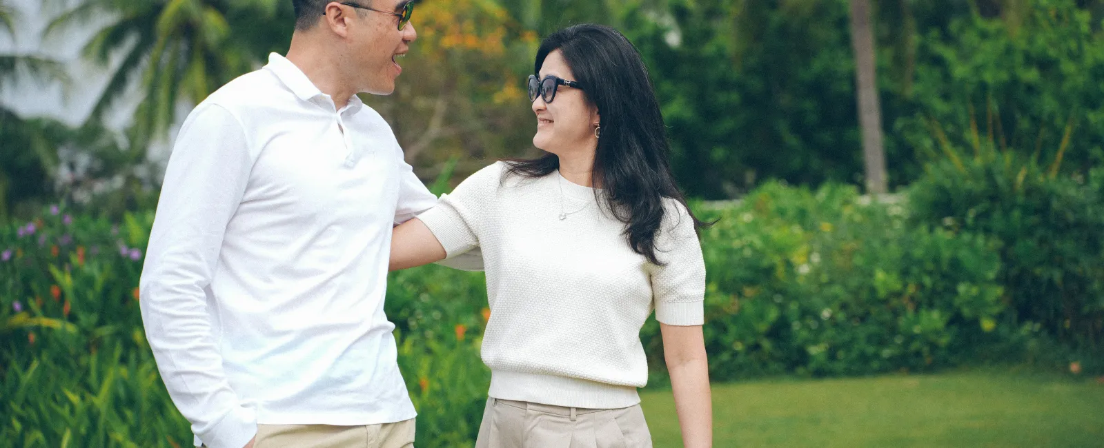 Couple wearing sunglasses and casual clothes enjoying a walk in a green garden with palm trees and buildings.