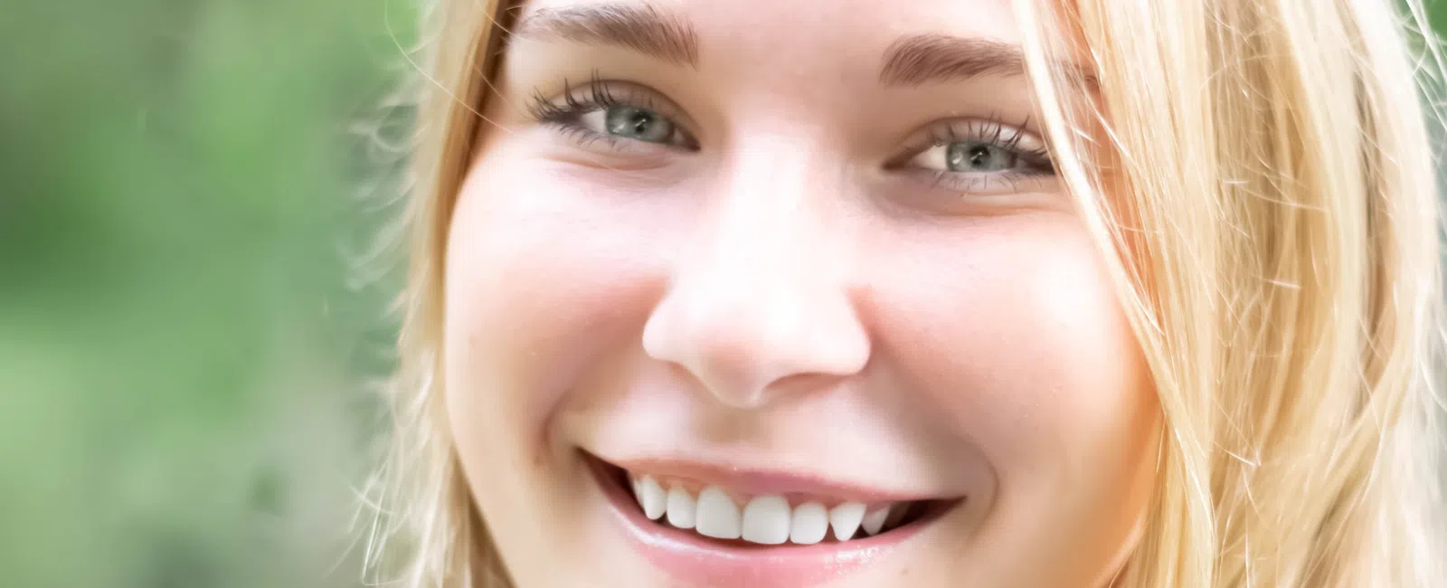 Close-up of a smiling young woman with blonde hair and blue eyes against a blurred green background