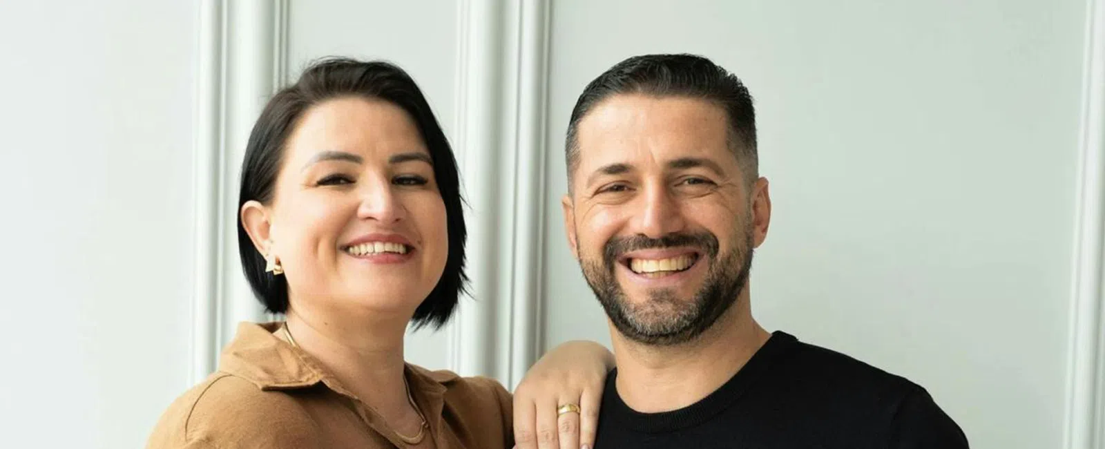 Smiling couple standing close with arm around shoulder against a light gray paneled wall background.