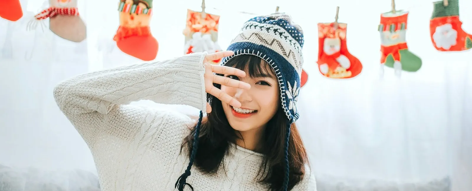 Smiling woman in cozy sweater and winter hat sitting on couch with festive Christmas stockings in background