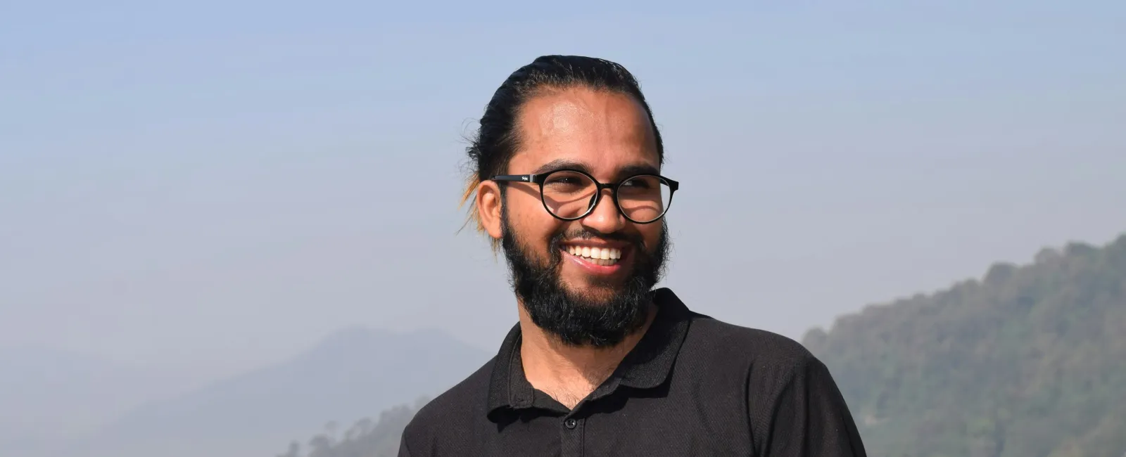 Smiling bearded man wearing glasses and black polo shirt outdoors with mountains and prayer flags in background