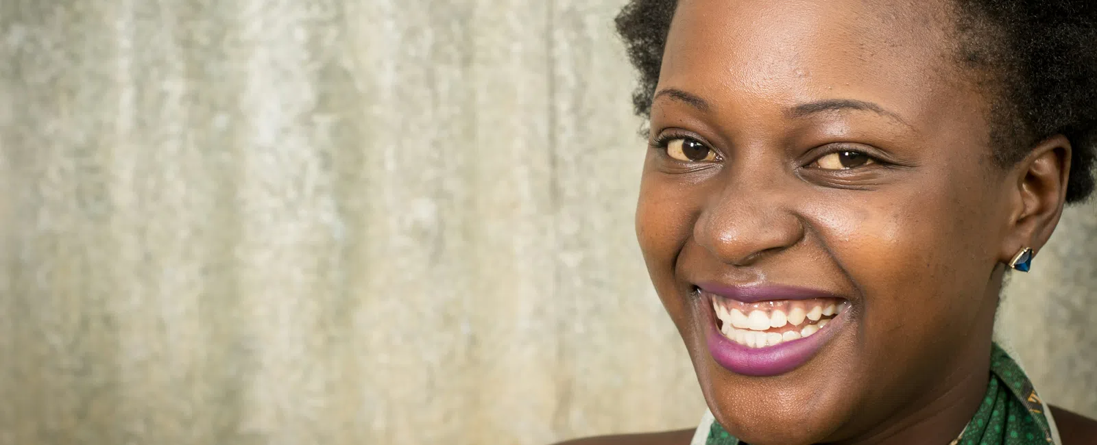 Smiling Black woman with short natural hair and vibrant purple lipstick wearing a green patterned top.