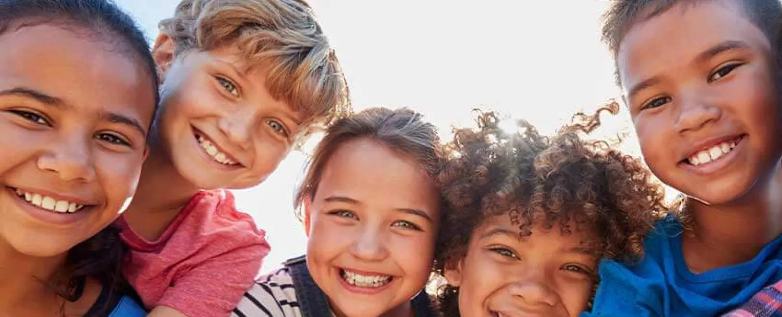 Five happy children smiling together outdoors against a bright blue sky.