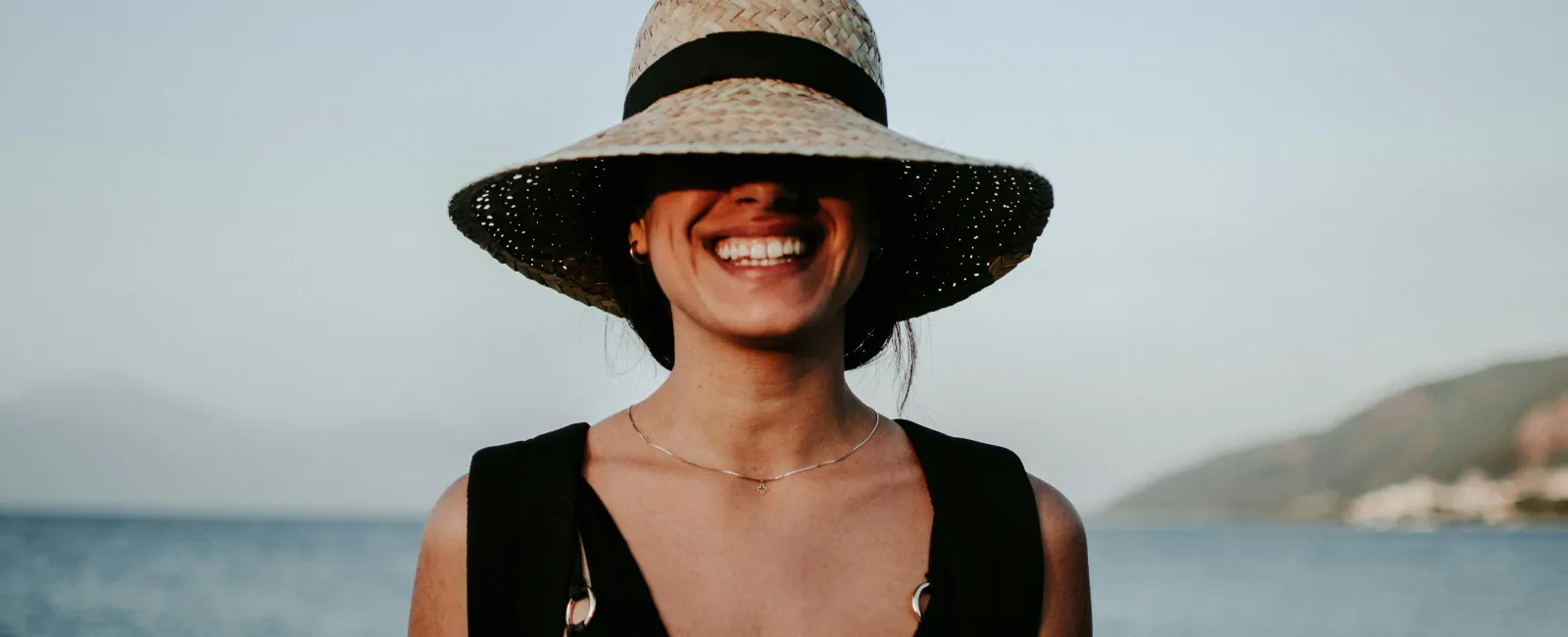 Smiling woman wearing a wide-brimmed hat by the beach during sunset.