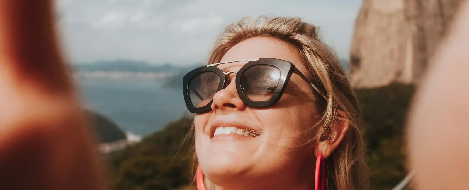 Smiling woman wearing sunglasses and pink earrings taking a selfie outdoors with a mountain and blue sky background