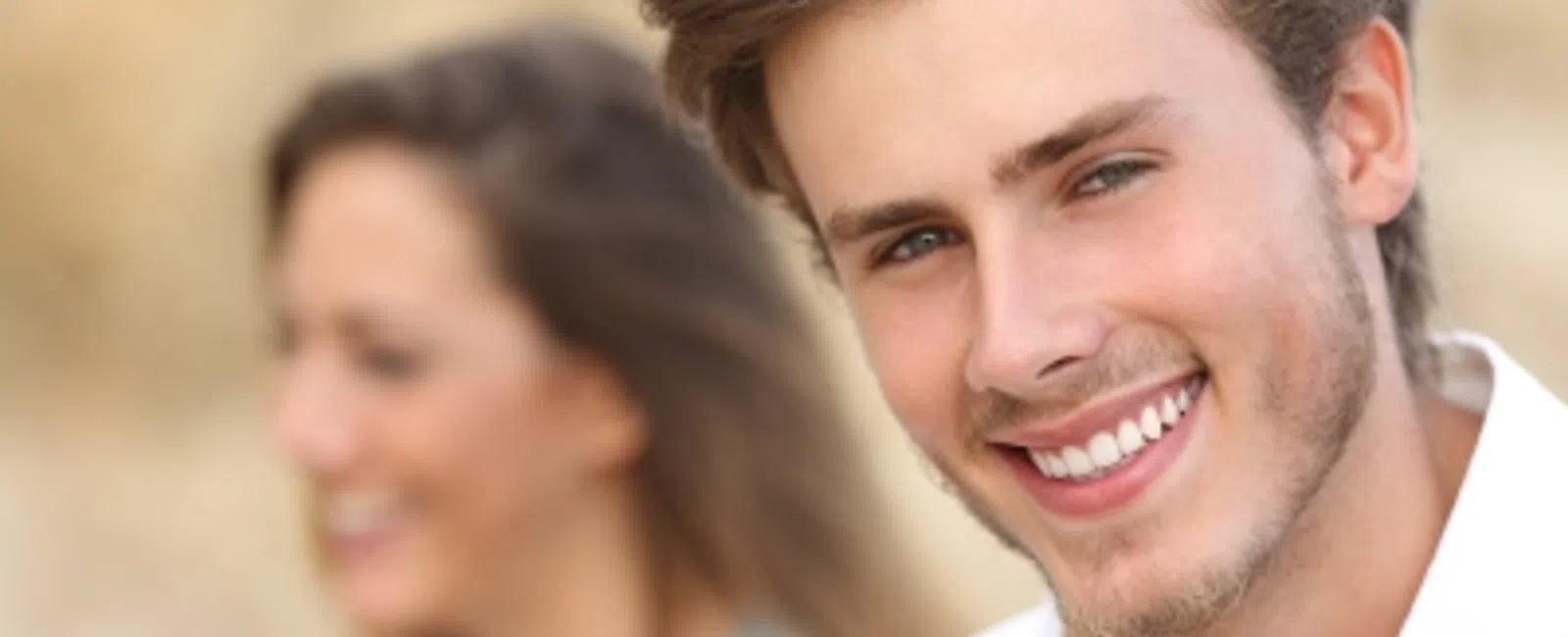 Smiling young man outdoors with a blurred woman in the background, showcasing joy and friendship.