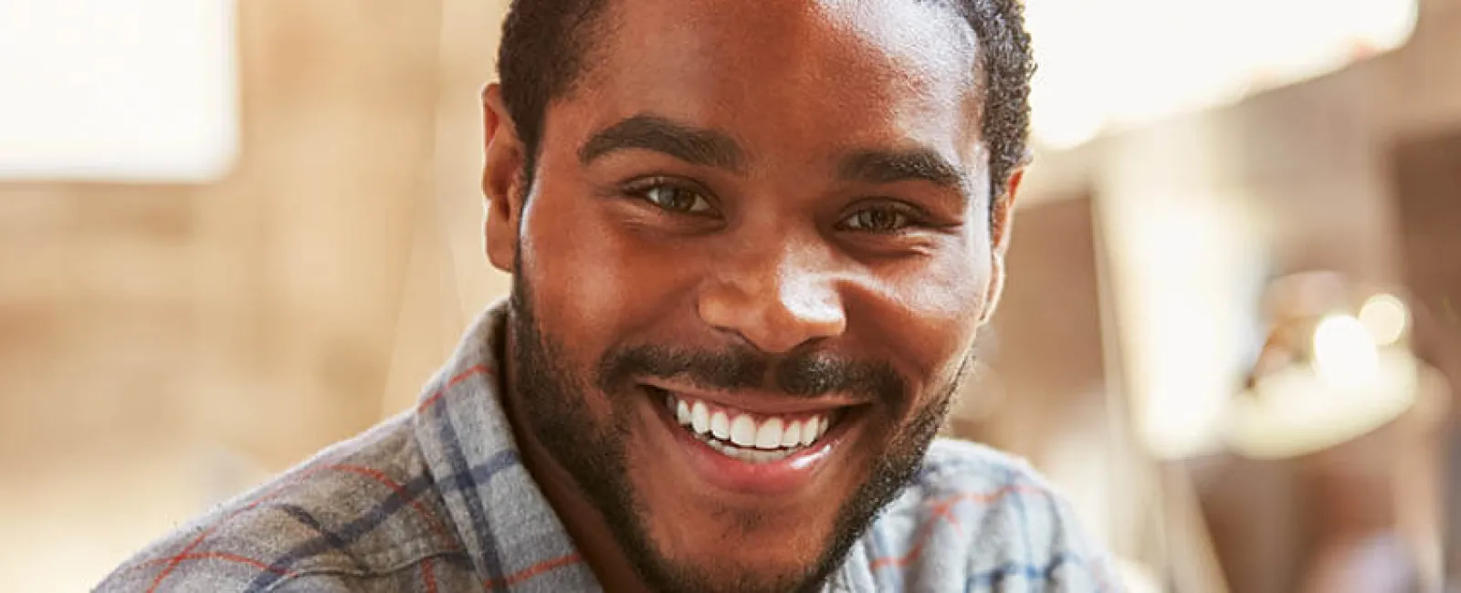 Smiling young man in a casual checkered shirt, seated in a bright modern workspace.