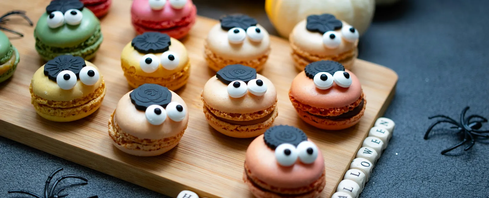 Halloween macarons with googly eyes and cookie bats on a wooden board with decorative spiders and pumpkin background.
