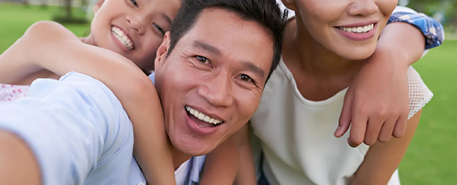 Happy Asian family of four smiling and embracing outdoors on a sunny day with green trees and lawn in background.