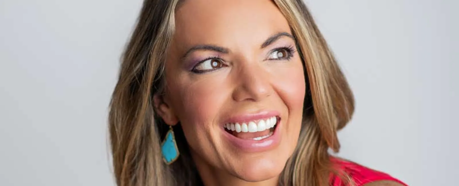Smiling woman with long blonde hair wearing a red top and turquoise earrings looking to the side.