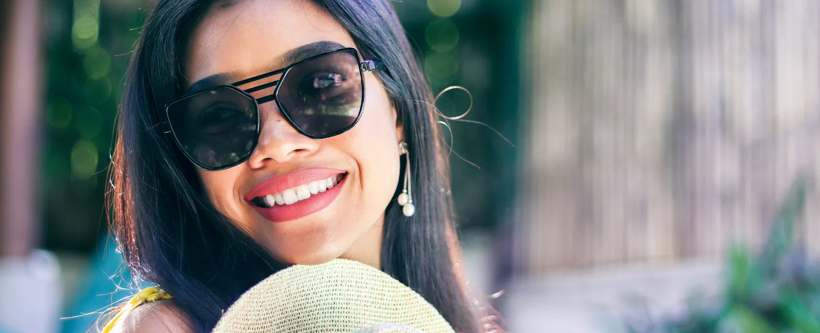 Smiling woman wearing sunglasses and holding a straw hat outdoors with blurred green and wooden background.