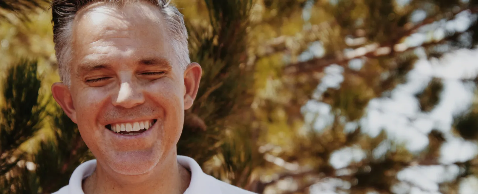 Smiling middle-aged man with gray hair in white polo shirt outdoors with sunlit pine trees in the background