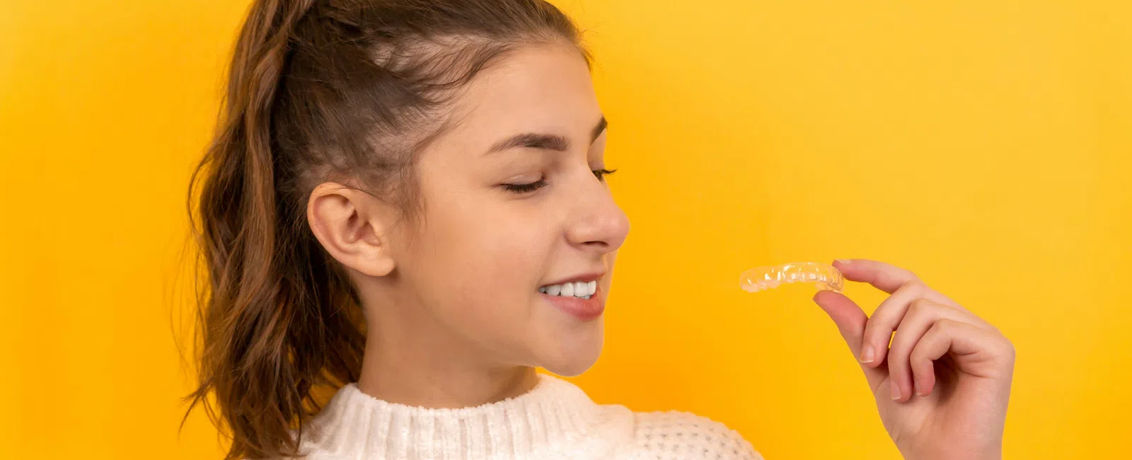 Young woman in a white sweater holding and smiling at a clear dental aligner against a yellow background.