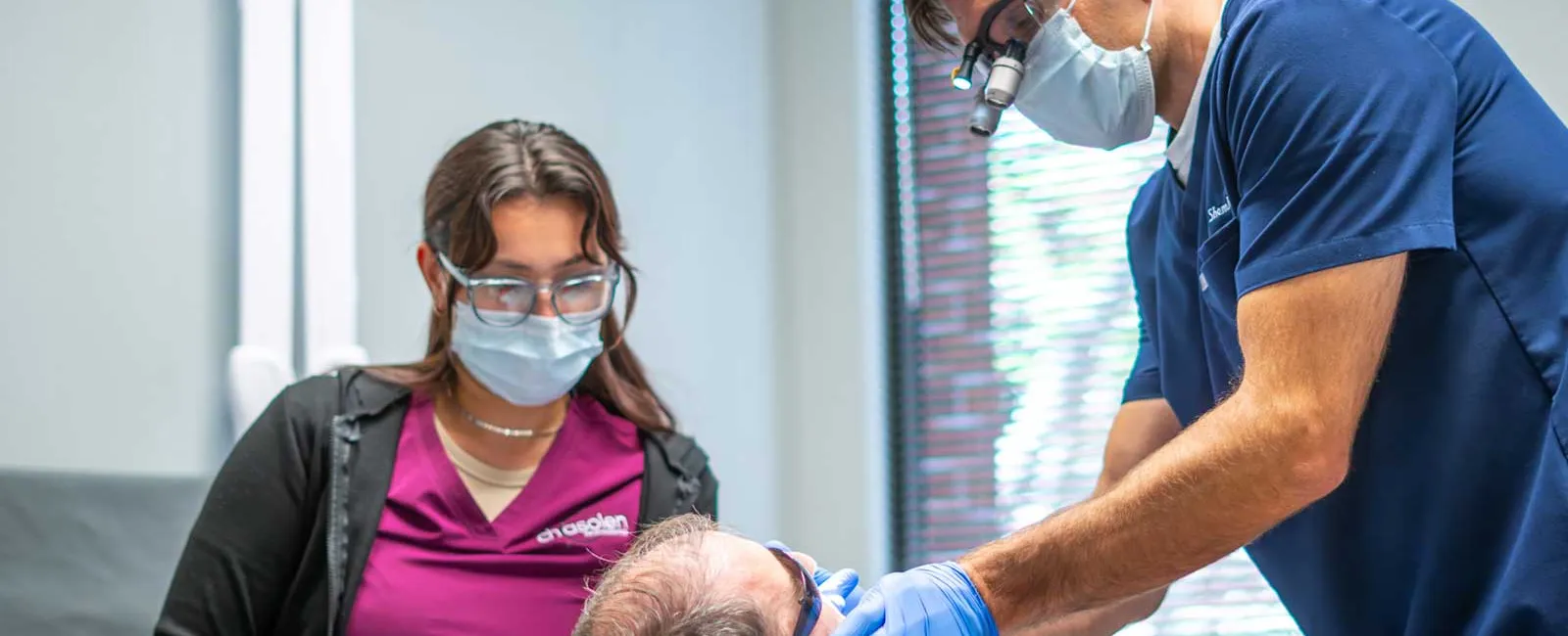 Dentist and assistant wearing masks and gloves treating a patient in a dental clinic with modern equipment.