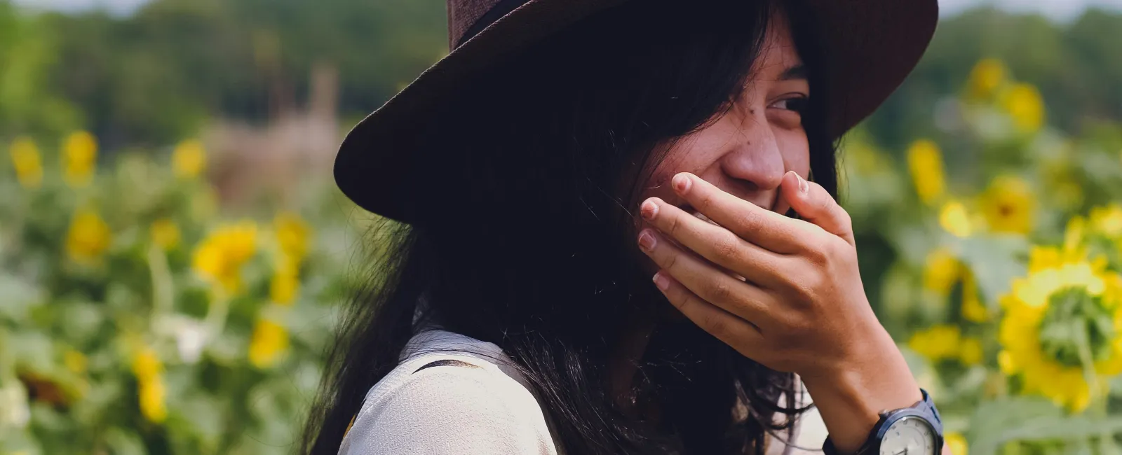 Woman laughing amidst vibrant sunflowers wearing a hat and a patterned shirt.