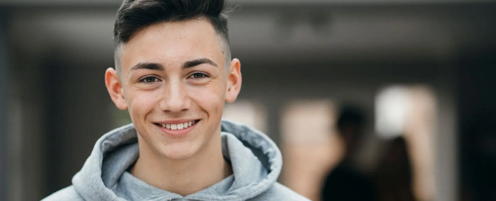 Smiling young man in a gray hoodie, indoors with a blurred background of people.