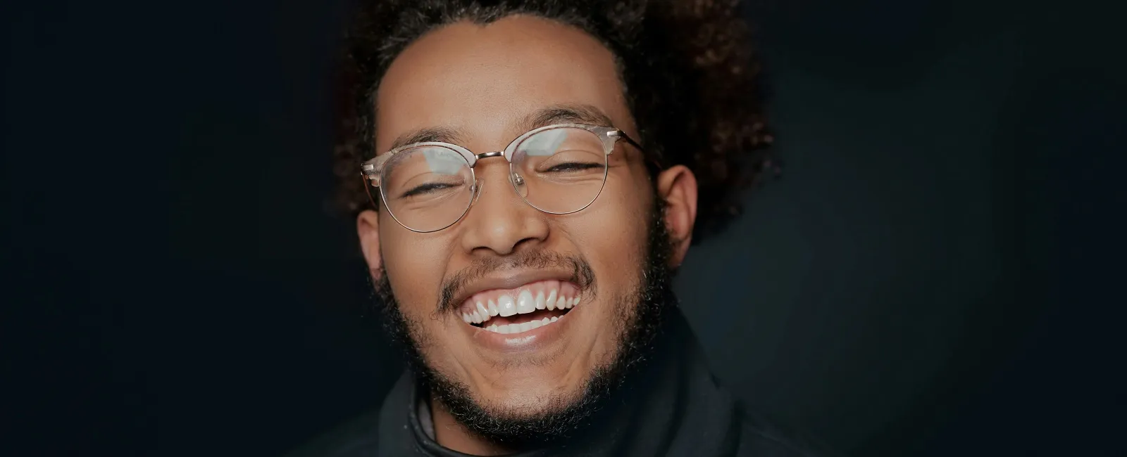 Happy young man with glasses and curly hair smiling against a dark background