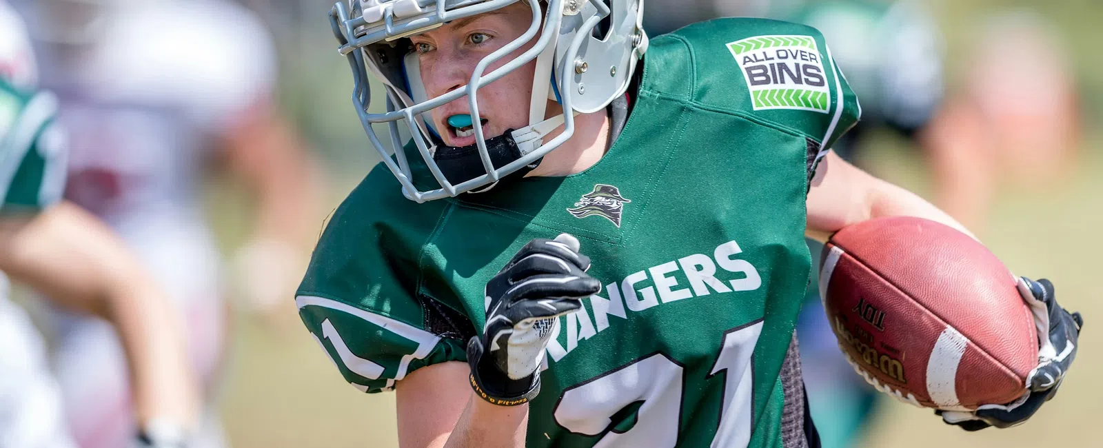 Youth football player in green Rangers jersey number 21 running with the ball during a game on a sunny day