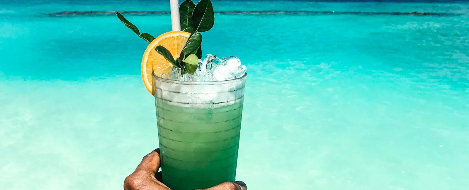 Hand holding a green cocktail with lemon garnish against turquoise ocean and yacht on horizon under blue sky.