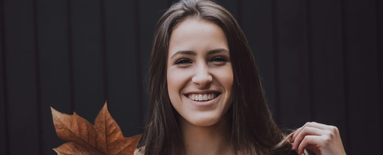 Smiling young woman holding a large brown autumn leaf against a dark wooden background