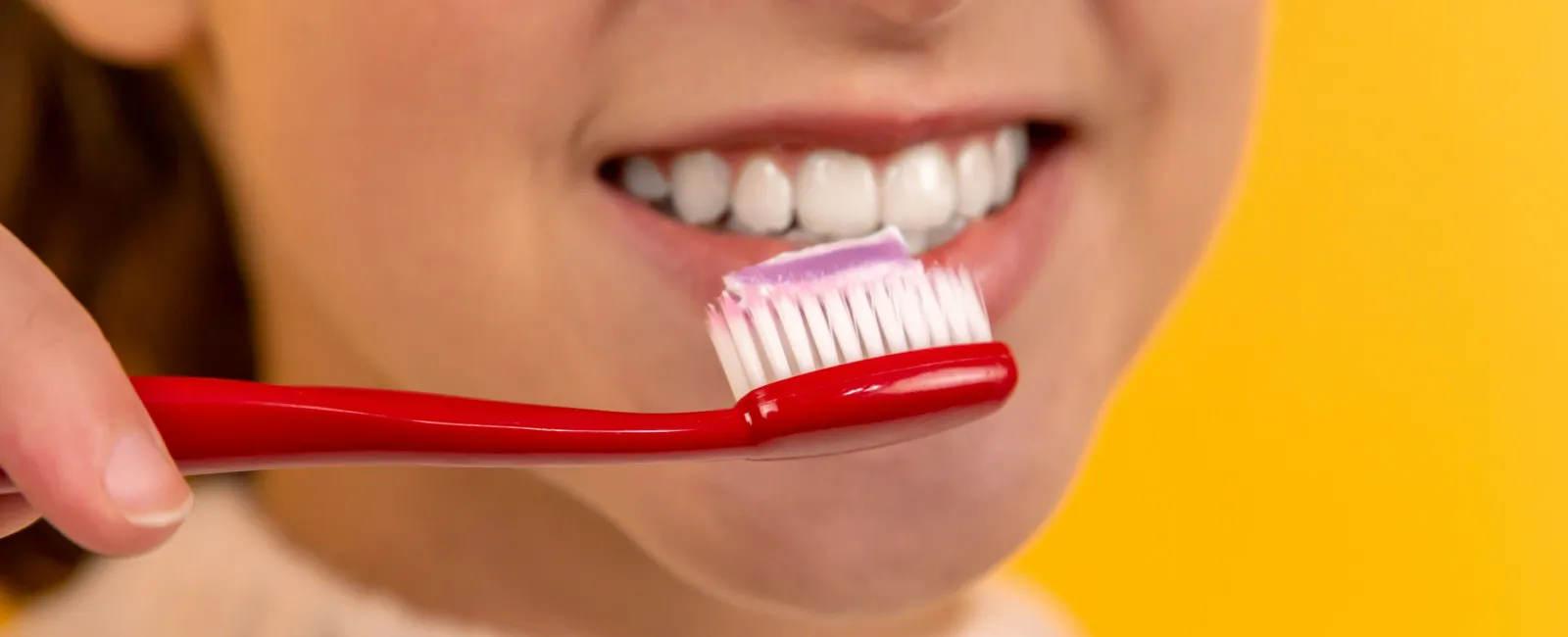 Close-up of a smiling person holding a red toothbrush with purple toothpaste against a yellow background
