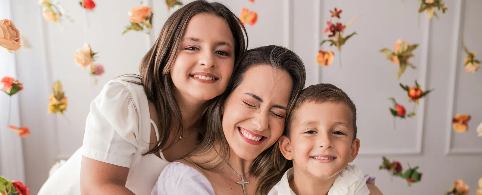 Smiling mother with daughter and son hugging amid hanging flowers in a bright, cheerful room.