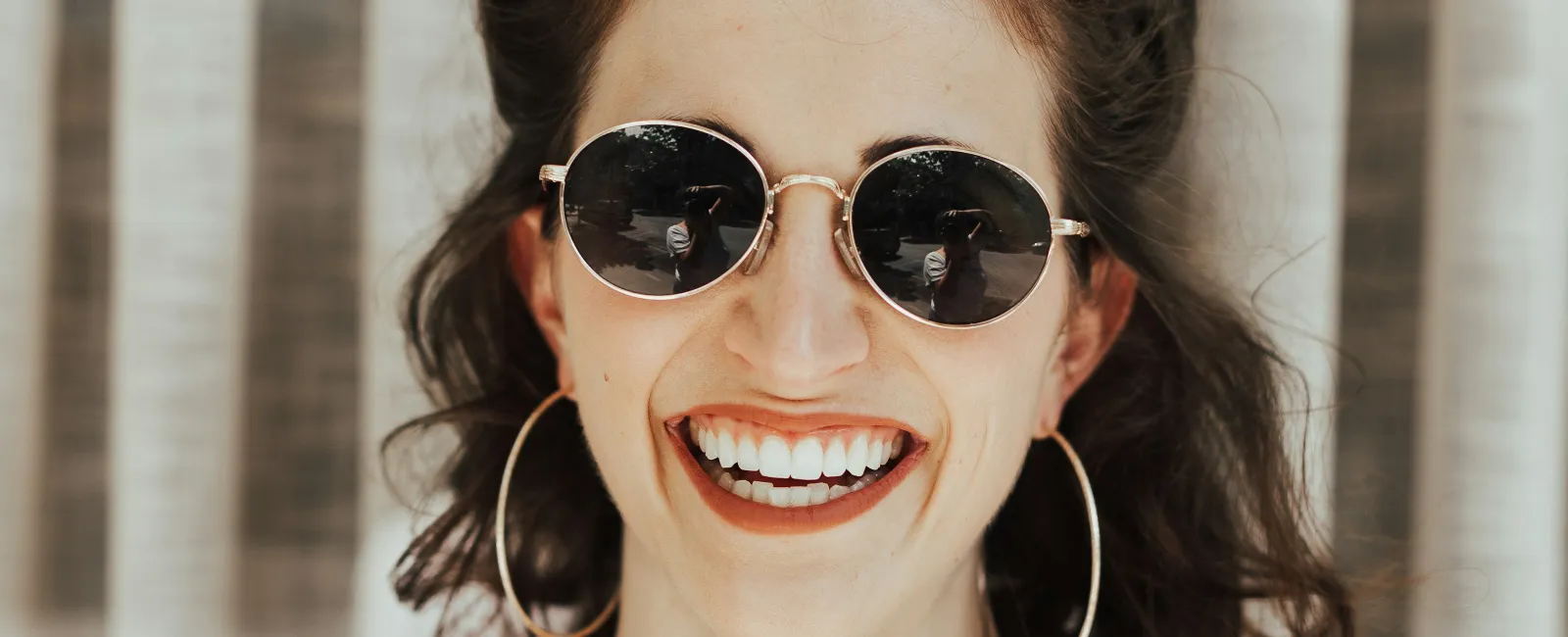 Smiling woman in round sunglasses and checkered overalls, posing against draped backdrop.