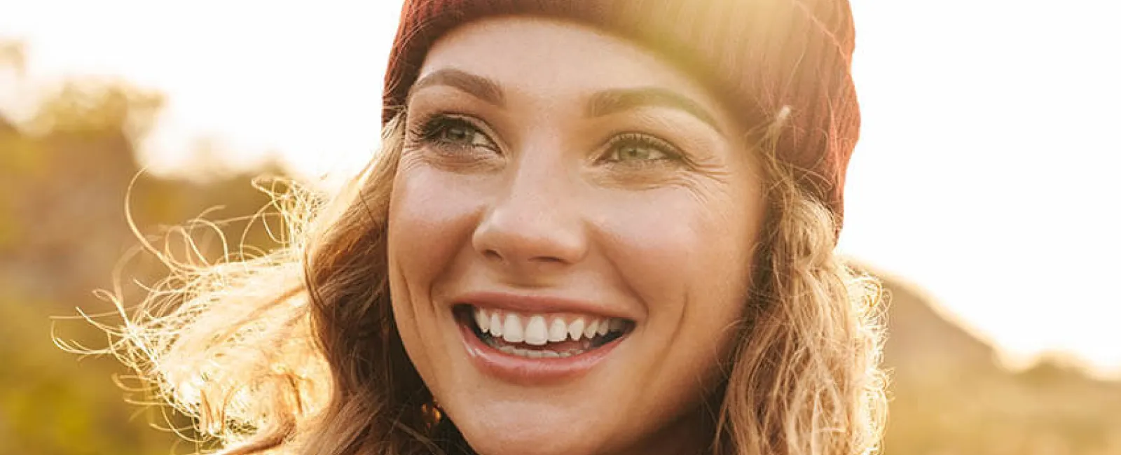 A smiling woman wearing a beanie and a green and white jacket, with sunlight glowing behind her.