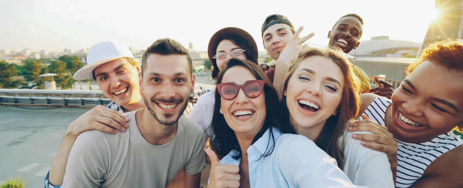 Group of diverse young adults smiling and taking a selfie outdoors on a sunny day at sunset.