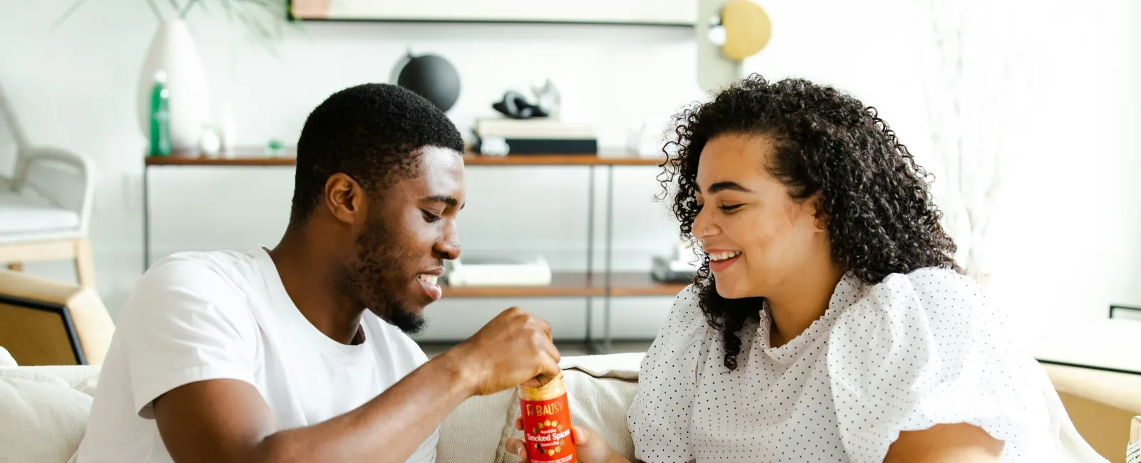 Couple smiling and sharing a bottle of hot sauce while sitting on a couch with snacks at home.
