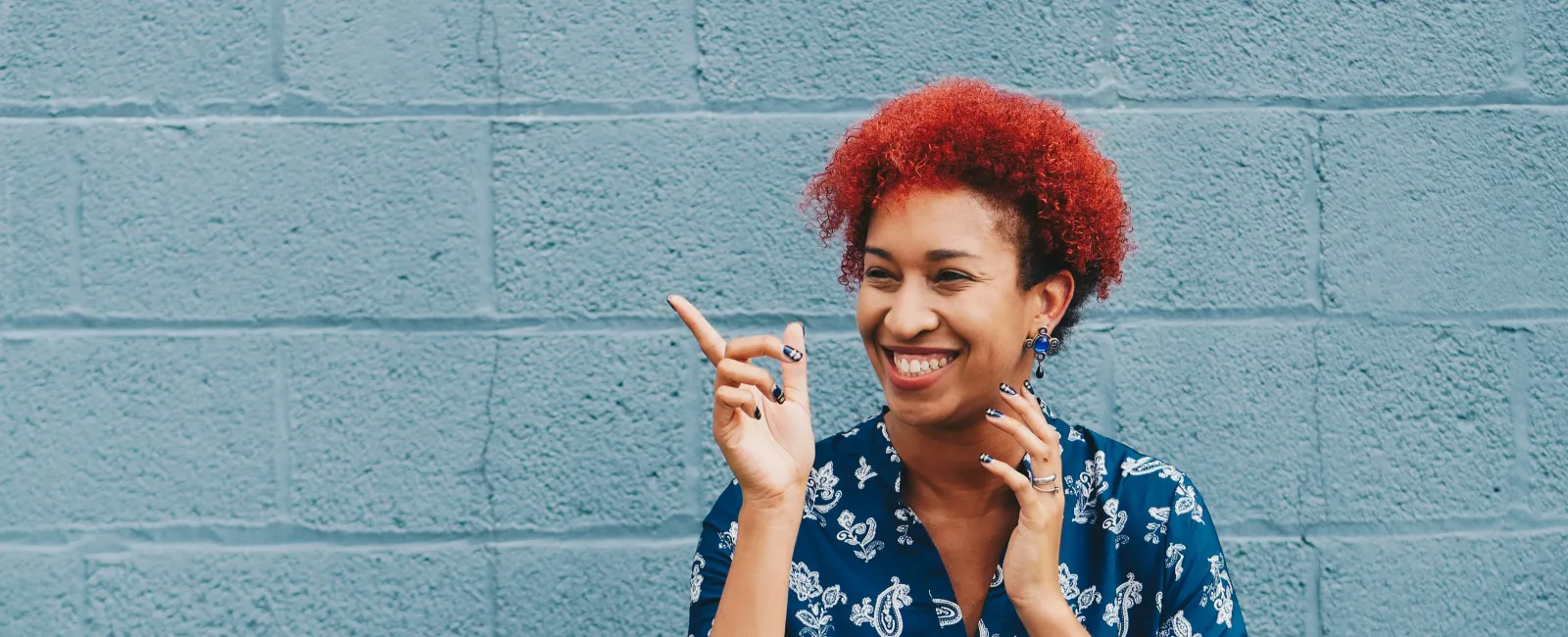 Smiling woman with red curly hair in a blue floral dress gestures happily against a gray brick wall background.