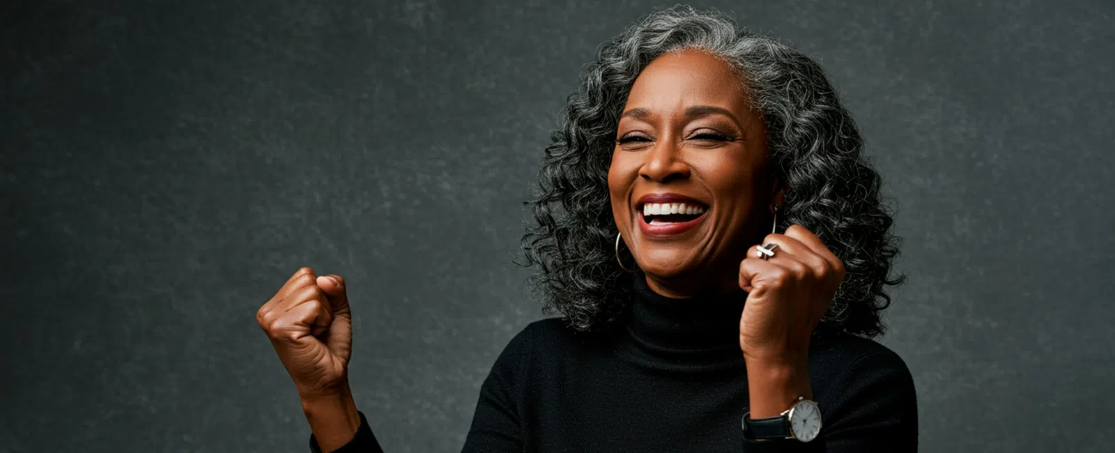 Happy African American woman with gray curly hair celebrating with clenched fists against a dark background.