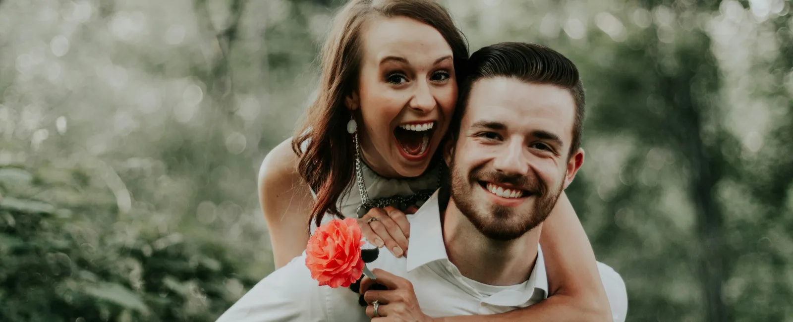 Happy couple outdoors with woman holding a flower on man's back, smiling in a green forest setting.
