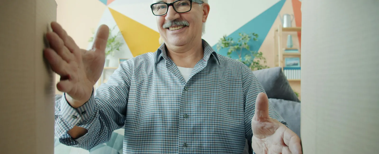 Smiling elderly man with glasses unpacking cardboard boxes in a brightly colored room.
