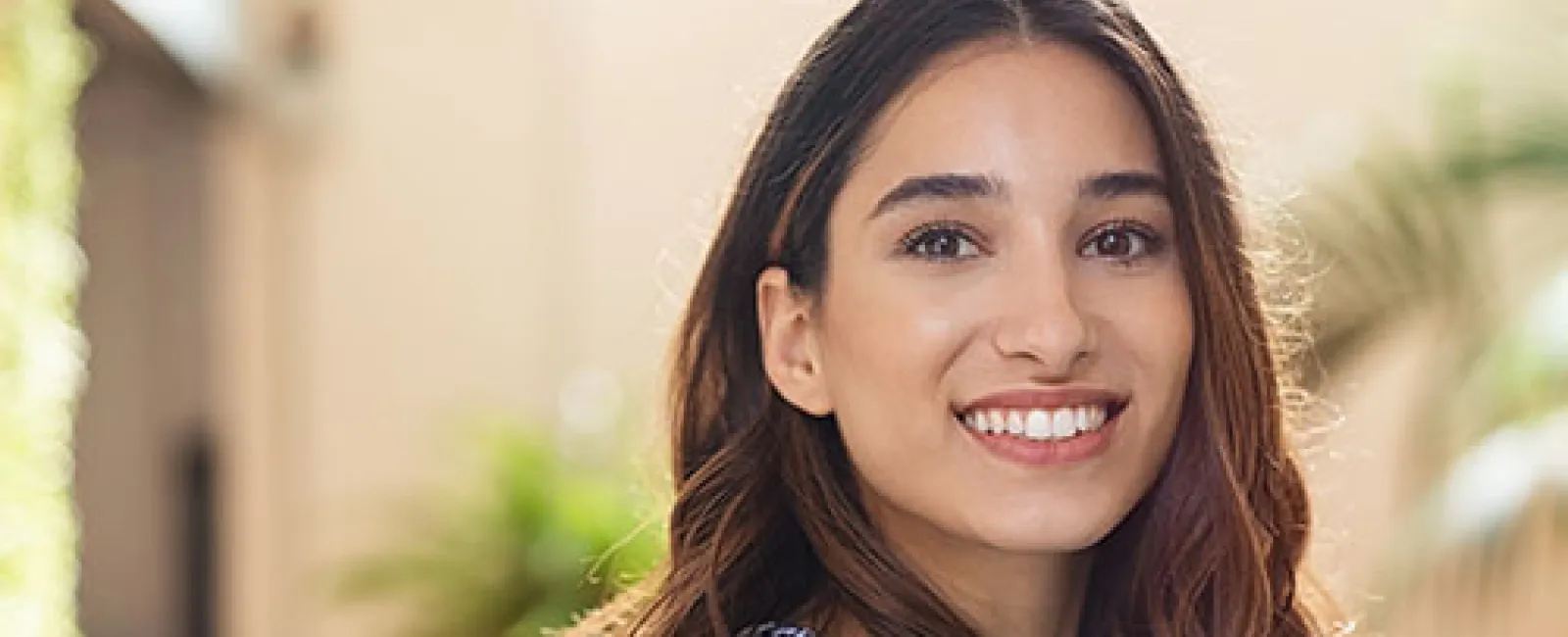 Smiling young woman with long brown hair wearing a floral blouse, sitting outdoors in soft natural light.