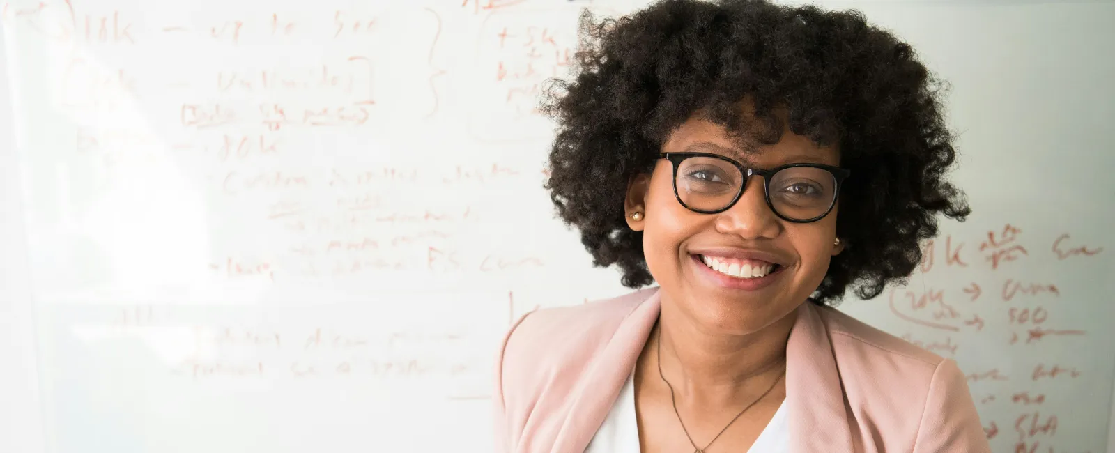 Smiling woman with glasses and curly hair standing in front of a whiteboard with notes and diagrams.