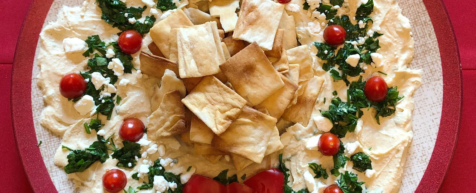Festive holiday hummus platter decorated with cherry tomatoes, parsley, pita chips, and red pepper bow on a plate.