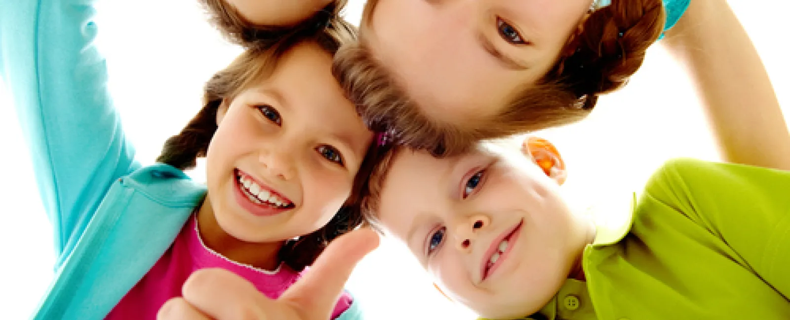 Four smiling children in colorful clothes huddled in a circle giving thumbs up and looking down at the camera.