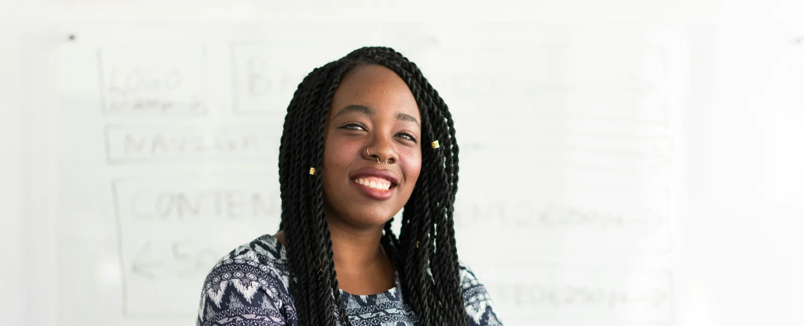 Confident Black woman with braided hair and patterned shirt stands with arms crossed in front of whiteboard