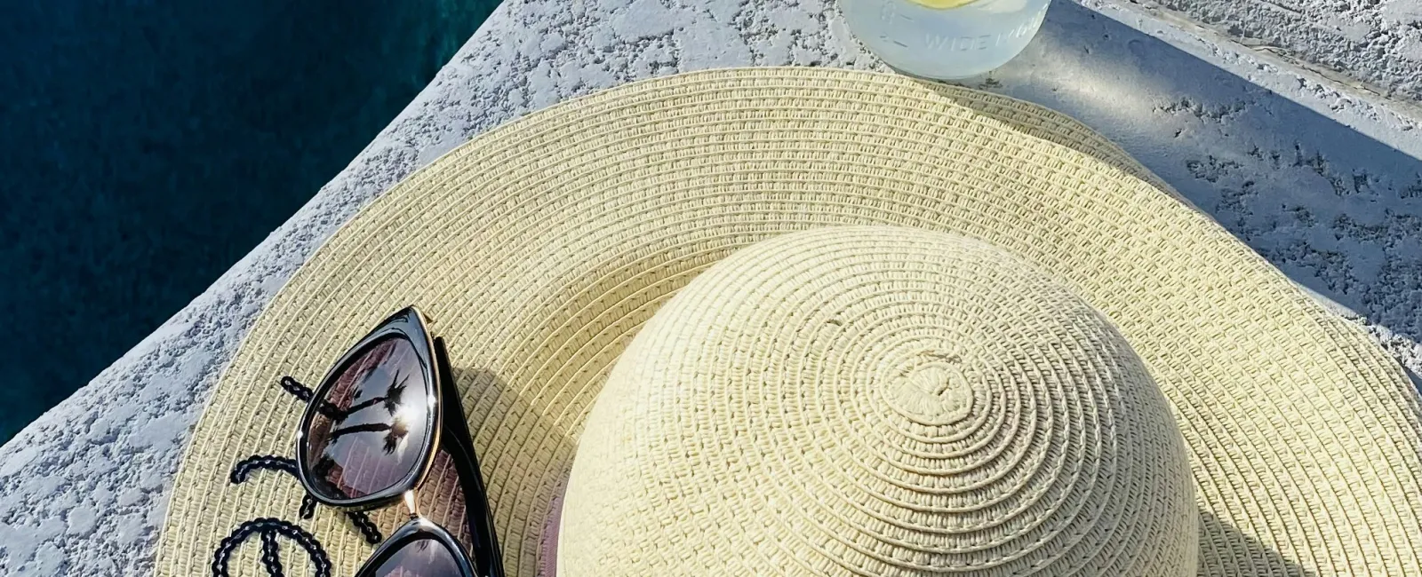 Straw sunhat with sunglasses by a poolside next to a cold drink with lemon and straw on hot sunny day.