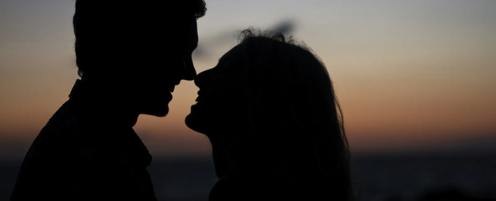 Silhouetted couple facing each other intimately during a sunset by the ocean with soft sky colors.