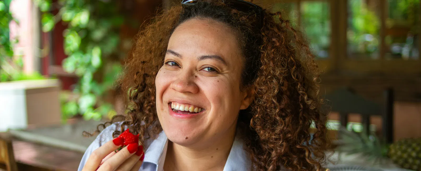 Smiling woman with curly hair eating fresh strawberries at a wooden table in a bright, cozy kitchen.